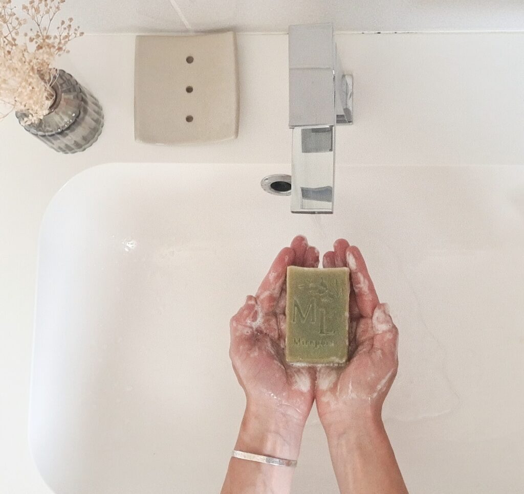 Porte savon en grés de couleur beige posé sur le bord d'un lavabo avec des mains qui portent un savon de couleur verte