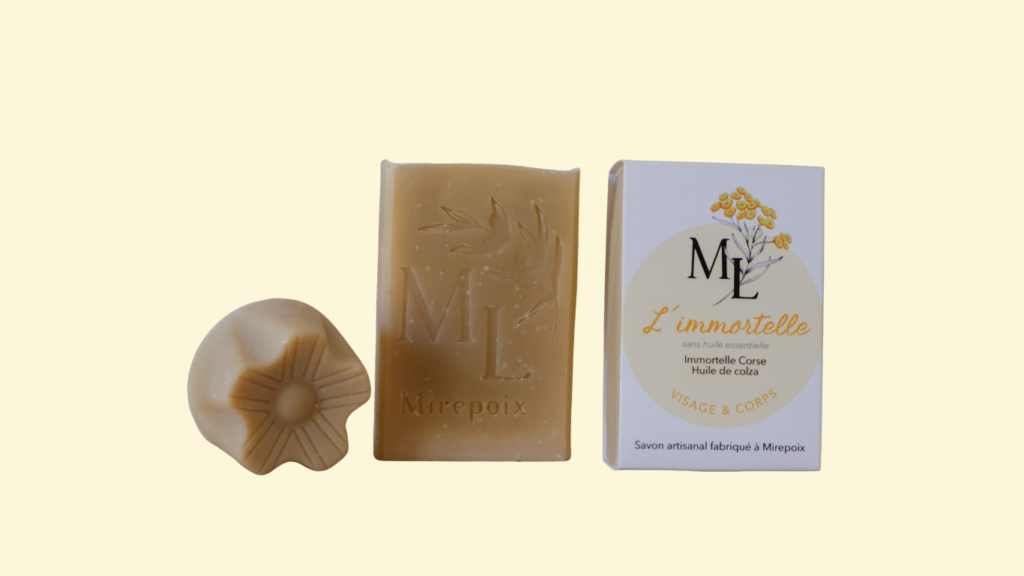 Savon rectangulaire de couleur jaune pâle posé verticalement à côté d'un autre savon avec le packaging illustré avec une fleur d'immortelle et une pastille jaune pastel et à côté d'un mini savon en forme de fleur