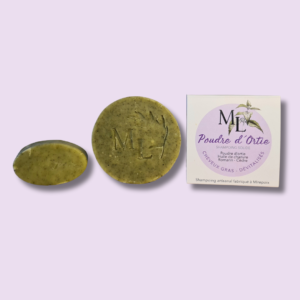 Shampoing de forme ronde de couleur vert posé à coté du shampoing avec son packaging de couleur mauve et une feuille d'ortie et un shampoing de forme ovale